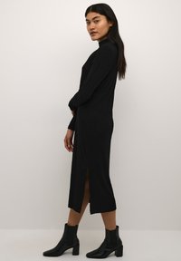 Schwarzes geripptes Strickkleid mit langen Ärmeln, hohem Kragen, seitlichem Schlitz und tailliertem Design. Das Model trägt schwarze Knöchelstiefel.