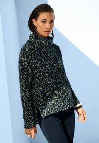 Frau trägt einen grob gestrickten Pullover mit grünen, blauen und schwarzen Garnen, schwarze Hosen und große schwarze geflochtene Ohrringe vor einer hellblauen Wand.