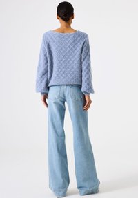 Femme avec un chignon portant un pull texturé bleu clair et un jean bleu clair à jambes larges, se tenant de dos sur un fond blanc.