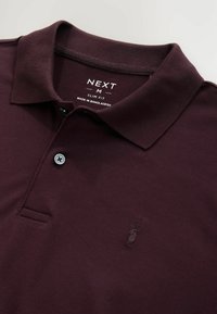 Tummanpunainen polo-paita slim fit -mallilla, jossa on röyhelökaulus, yksittäinen nappi ja huomaamaton brodeerattu logo rinnassa.