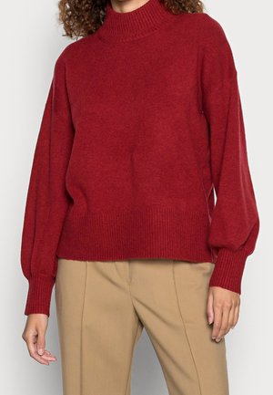 Personne portant un pull en tricot rouge ample avec des manches bouffantes et un pantalon beige, visible des épaules aux hanches sur un fond uni.