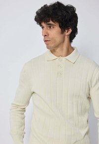Homme aux cheveux noirs frisés portant un polo à manches longues côtelé crème, regardant vers la gauche devant un fond uni.