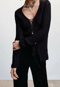 Czarny, dziany kardigan z dekoltem w serek, szczegółami sznurowania z przodu, prążkowaną fakturą i długimi rękawami. Połączony z ciemnymi, aksamitnymi spodniami.