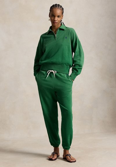 Femme aux cheveux tressés portant un sweat à capuche vert zippé et un jogging assorti avec un cordon blanc, debout les mains dans les poches.