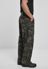 Pantalones cargo en un patrón de camuflaje oscuro, hechos de una tela resistente. Dos grandes bolsillos laterales, corte holgado y combinados con calzado negro.