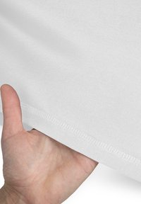 Ein weißer Stoff, der mit einer Hand, die die Kante hält, präsentiert wird, hat eine glatte Textur und sichtbare Nähte am Saum.