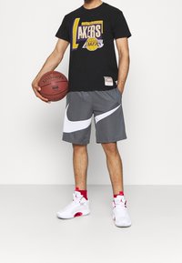 Maglietta nera con grafica "Los Angeles Lakers", pantaloni Nike grigi con logo bianco, scarpe da ginnastica rosse e bianche, mentre tiene un pallone da basket.