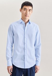 Camicia a maniche lunghe azzurro chiaro con collo button-down, caratterizzata da una texture liscia e bottoni a contrasto, indossata con pantaloni scuri.