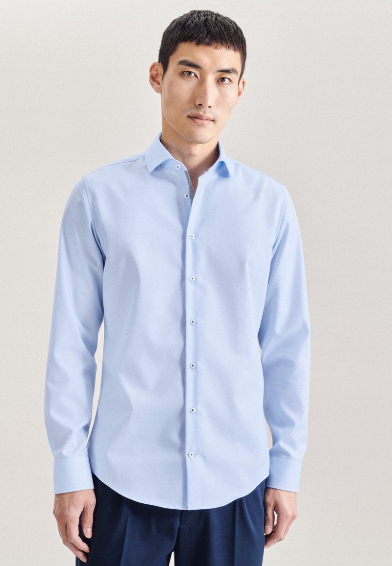 Camicia a maniche lunghe azzurro chiaro con collo button-down, caratterizzata da una texture liscia e bottoni a contrasto, indossata con pantaloni scuri.