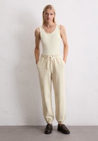 Helles gelbes ärmelloses Bodysuit kombiniert mit beigen Sweatpants, die über einen elastischen Bund und einen Kordelzug verfügen; das Model trägt dunkle Schuhe.