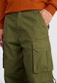 Oliwkowe spodnie cargo wykonane z teksturowanej tkaniny, z bocznymi kieszeniami i zapięciem na guzik, prezentowane z pomarańczową koszulą z długim rękawem.