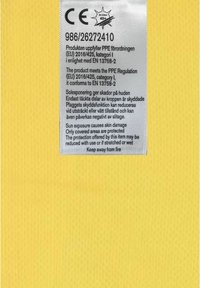 Vit vårdtagg på en gul texturerad tygbakgrund. Taggen inkluderar CE-märkning, säkerhetsinstruktioner och överensstämmelseinformation för PPE-föreskrifter.