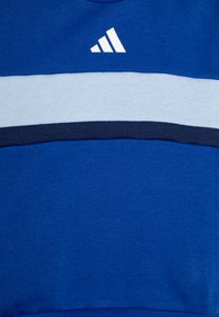 Sudadera azul con una franja horizontal en azul claro y azul marino. Logo blanco de Adidas en la parte superior izquierda. Tejido suave y texturizado.
