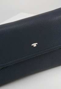 Granatowy, teksturowany portfel skórzany z małym, srebrnym emblematem umieszczonym na przednim klapce, położony na białej powierzchni.