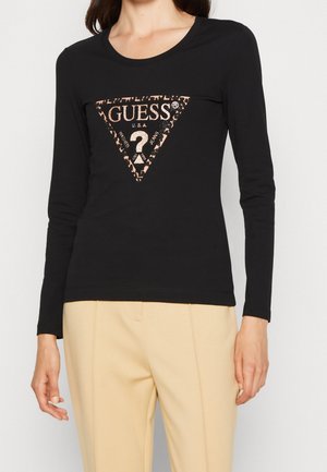 Femme portant un t-shirt noir à manches longues avec un logo Guess en triangle imprimé léopard et un pantalon beige, debout sur un fond blanc.
