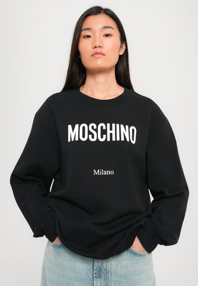 Sudadera negra de Moschino con logo blanco, de corte holgado, cuello redondo y puños acanalados, combinada con jeans de mezclilla azul claro.