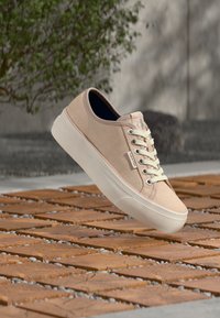 Μπεζ αθλητικό παπούτσι Calvin Klein με λευκά κορδόνια που αιωρείται πάνω από ξύλινα πλακάκια σε εξωτερικό χώρο, με πράσινο θάμνο και γκρι τοίχο στο φόντο.