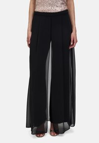 Pantalons larges noirs avec superposition transparente, design plissé et taille élastique. Associé à un haut pailleté et des talons métalliques.