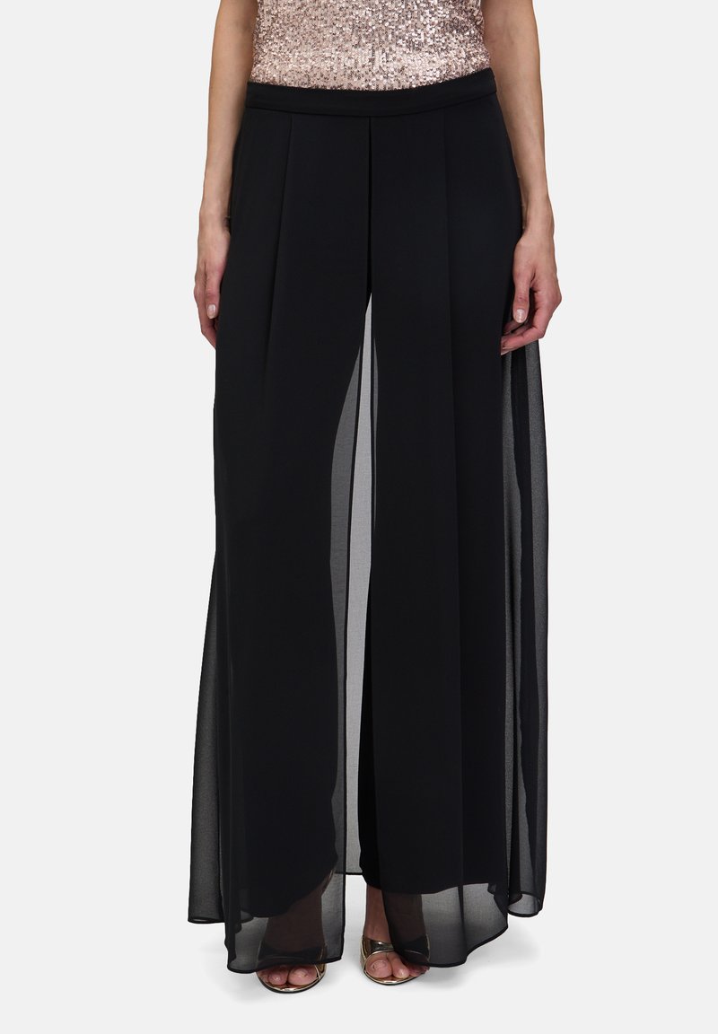 Pantalons larges noirs avec superposition transparente, design plissé et taille élastique. Associé à un haut pailleté et des talons métalliques.