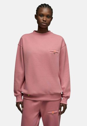 Une personne portant un ensemble ample rose composé d'un sweat-shirt et d'un pantalon assortis, avec de petites poches boutonnées sur la poitrine et la cuisse, debout devant un fond uni.