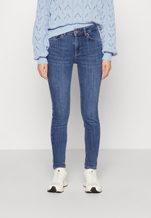 Kvinde iført lyseblå striktrøje, tætsiddende blå jeans og hvide sneakers står mod en ensfarvet lys baggrund.