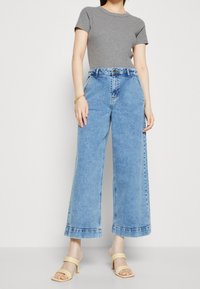 Nainen, jolla on yllä vaaleansiniset leveälahkeiset farkut, harmaa lyhythihainen paita, beigellä korolla varustetut sandaalit ja kultaketju, seisoo vaaleaa taustaa vasten.