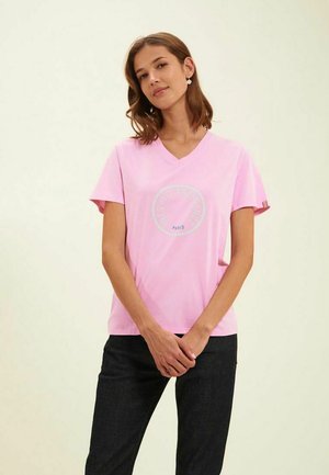 T-shirt en coton rose avec un col en V et des manches courtes. Présente un imprimé graphique circulaire indiquant "Paris" au centre. Pantalons sombres.