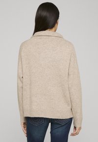 Pull en tricot beige avec col châle, ourlet côtelé et manches larges. Le dos présente une silhouette décontractée et carrée ainsi qu'une texture douce.
