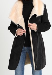 Cappotto in suede nero con colletto in pelliccia sintetica beige, chiusura con zip frontale, maniche lunghe e design di lunghezza media. Presenta una fodera morbida e testurizzata.