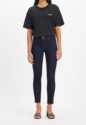 Kobieta ubrana w czarny t-shirt Levi's, ciemnoniebieskie, taliowane jeansy z wysokim stanem oraz czarne sandały na słupku z paskami, stojąca na białym tle.