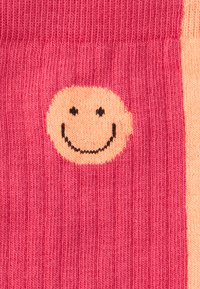 Șosete tricotate în tonuri de roz și piersică, cu un grafic mare al feței zâmbitoare în detalii negre, evidențiind textura răsucită.