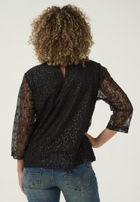 Top in pizzo nero con maniche a tre quarti, caratterizzato da un dettaglio a chiave sul retro e un motivo ondulato su tutta la superficie. Abbinato a jeans in denim blu.