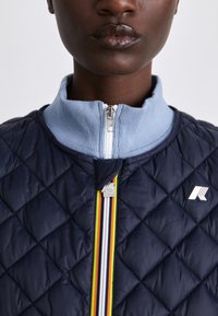 Quilted navy jacket with a light blue ribbed collar and multi-colored zipper detail, featuring a silver logo on the front.