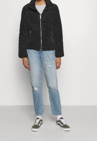 Svart kordjacka med dragkedja framtill, mjuk textur och hög krage. I kombination med ljusblå slitna jeans och gröna skor med vita detaljer.