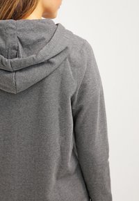 Femme portant un sweat à capuche gris uni, vue de dos sur un fond clair uni.