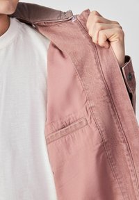 Personne portant une chemise blanche tient une veste rose ouverte pour révéler la poche intérieure et les détails du tissu.