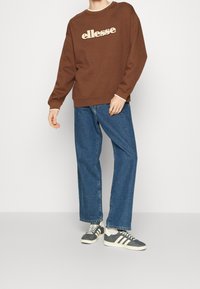 Hnědá mikina s texturovaným krémovým logem, spárovaná s modrými širokými džíny a šedými teniskami s bílými pruhy a tkaničkami.