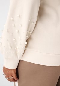 Crème trui met parelversieringen op de mouwen, geribbelde zoom en een gladde textuur. Gecombineerd met lichtbruine slim fit broek.