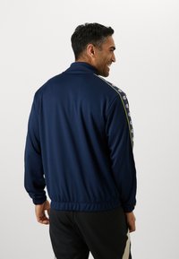 Veste bleu marine avec un col montant, dotée d'accents de logo le long des épaules et de poignets élastiques. Tissu lisse et léger.