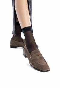 Mocasines de ante marrón con una suela plana negra, que presentan un diseño cosido y acentos metálicos, combinados con calcetines negros transparentes hasta el tobillo.
