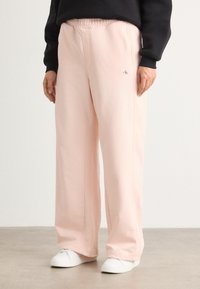 Pantalons de jogging rose pâle avec une taille élastique, des coutures latérales et un petit logo. Associés à des baskets blanches sur un sol neutre.