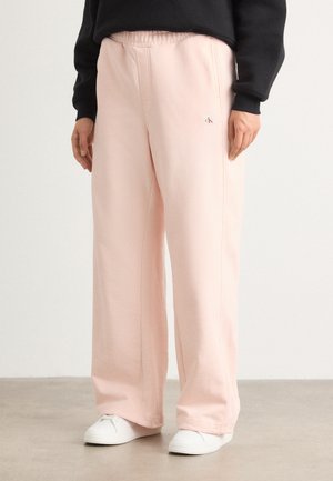 Lichtroze joggingsbroek met een elastische tailleband, zijnaad en een kleine logopatch. Gecombineerd met witte sneakers op een neutrale vloer.
