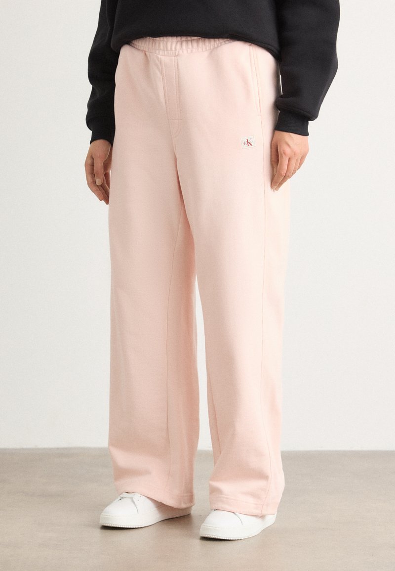 Pantalons de jogging rose pâle avec une taille élastique, des coutures latérales et un petit logo. Associés à des baskets blanches sur un sol neutre.