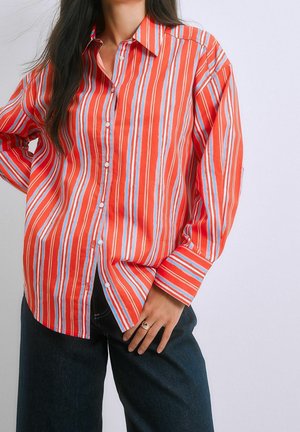 Femme portant une chemise rouge ample à boutons avec des rayures verticales blanches et bleu clair et un jean bleu foncé, main posée sur la hanche.