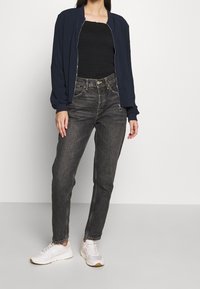 Kvinna i en marinblå zip-up-jacka, svart texturerad topp, högmidjade urtvättade svarta jeans och vita sneakers som står mot en vit bakgrund.