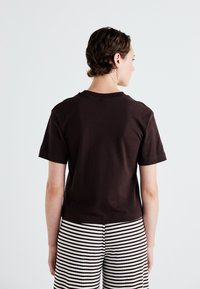 Brown short-sleeve t-shirt made of cotton, seen from the back, paired with black and white striped pants; smooth texture, standard crew neck.