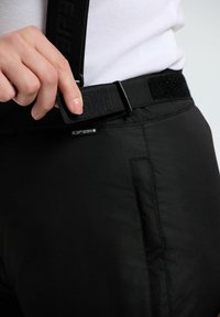 Ręka dostosowująca czarny pas z rzepem w czarnych spodniach z metką "Icepeak", noszonych z białą koszulą.