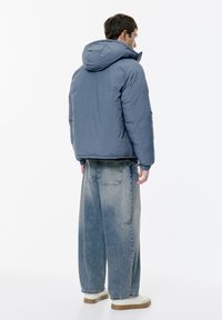 Man med ryggen mot kameran, iklädd en blå huva-puffjacka, vida bleknade blå jeans och vita sneakers med gummisulor.