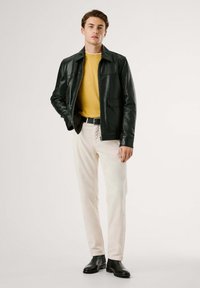 Chaqueta de cuero negra, suéter amarillo mostaza, pantalones de pana blancos, cinturón negro, botas negras tipo ankle, pose de pie, fondo liso.