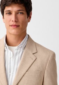Blazer beige claro con una tela texturizada y un bolsillo en el pecho, superpuesto a una camisa a rayas blanca y azul claro.